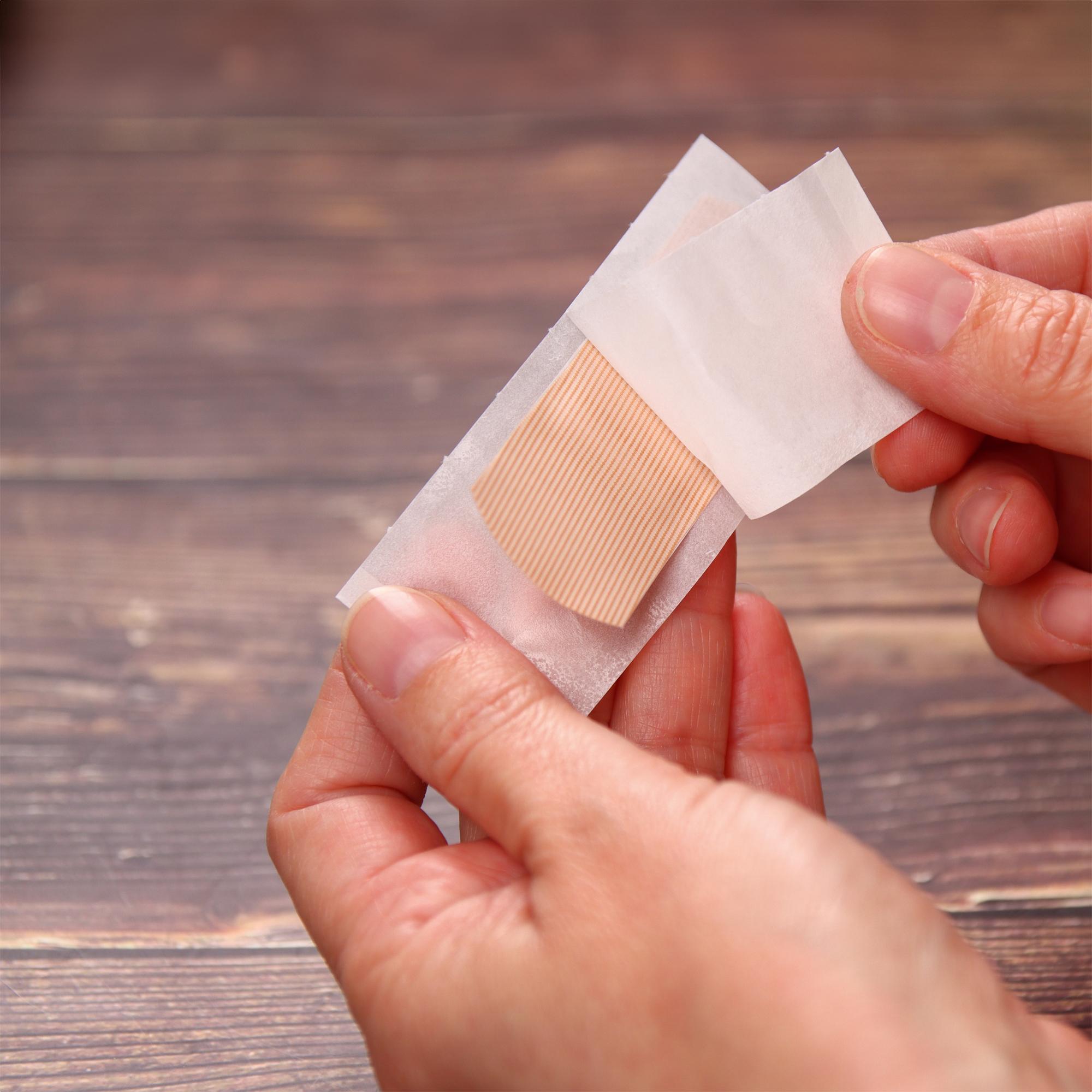 Ein Weroplast® AquaFlex Pflaster - Mix DIN 13169 in beige wird von zwei Händen gehalten, das flexible, wasserdichte Pflaster wird leicht angehoben.