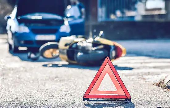 Gelbes und schwarzes Motorrad liegt umgekippt neben Warnschild auf Asphalt, Vorderreifen verdreht, kaputtes Auto mit offener Motorhaube im Hintergrund.