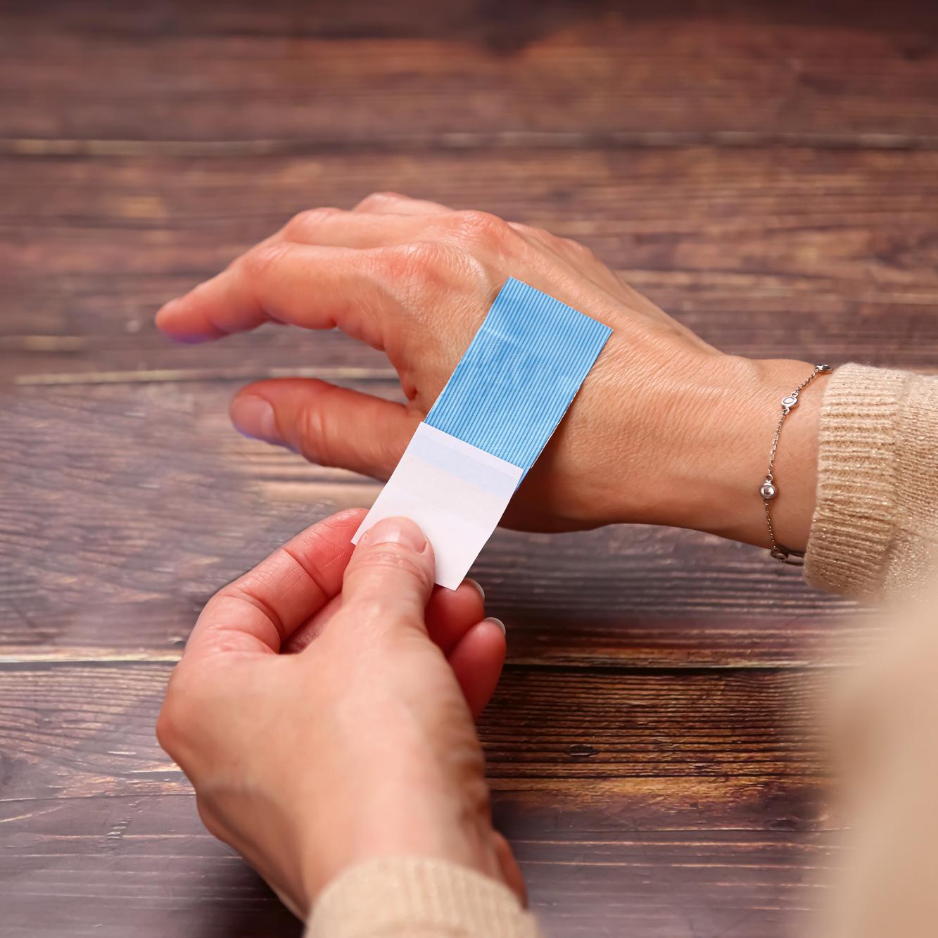 Eine Hand legt einen Weroplast® AquaFlex Detect Wundschnellverband mit wasserfester, blauer PU-Trägerfolie und Aluminiumdetektion am Handgelenk an.