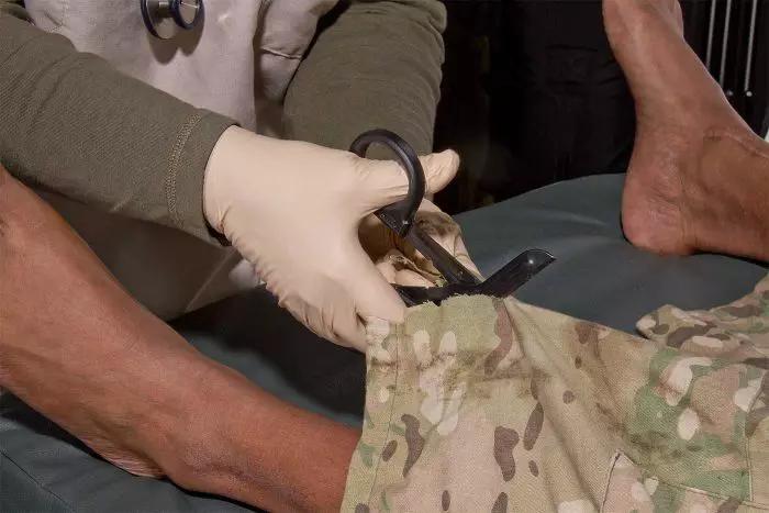 Ein Arzt mit Handschuhen schneidet mit einer NAR Trauma Shear - 15,8 cm eine tarnfarbene Verbandbinde vom Bein eines Patienten auf einer Trage.