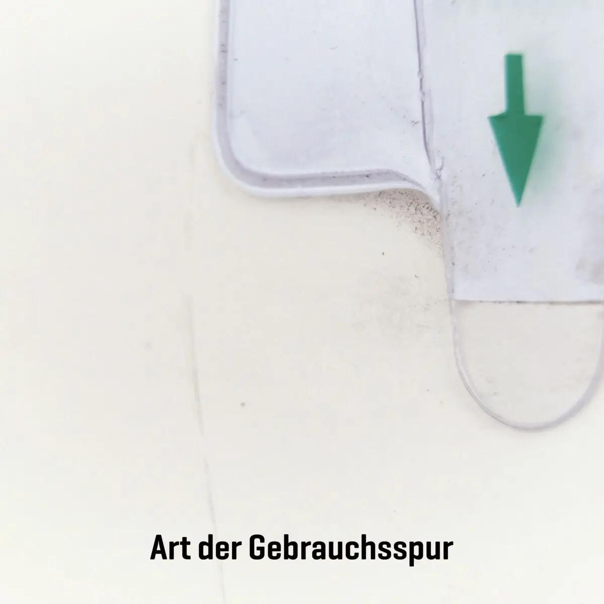 Ein weißes Kunststoffteil mit grünem Abwärtspfeil zeigt eine typische Gebrauchsspur des AED-Wandschrank (gebraucht) - ohne Alarmfunktion.