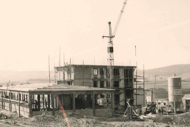 Schwarzweißfoto einer belebten Baustelle mit Arbeitern, eingerüstetem Rohbau, Kran, Baumaterial und Wasserturm in ländlicher Umgebung.