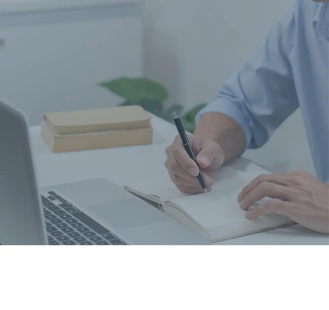 Eine Person in blauem Hemd schreibt mit schwarzem Stift in ein Notizbuch an einem weißen Schreibtisch mit Laptop und Büchern.