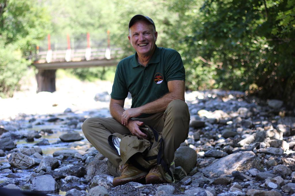 Mann im grünen Polo und Basecap sitzt lächelnd auf einer Steinbank am Fluss, neben ihm eine schwarze Tasche, im Hintergrund eine Brücke und Bäume.