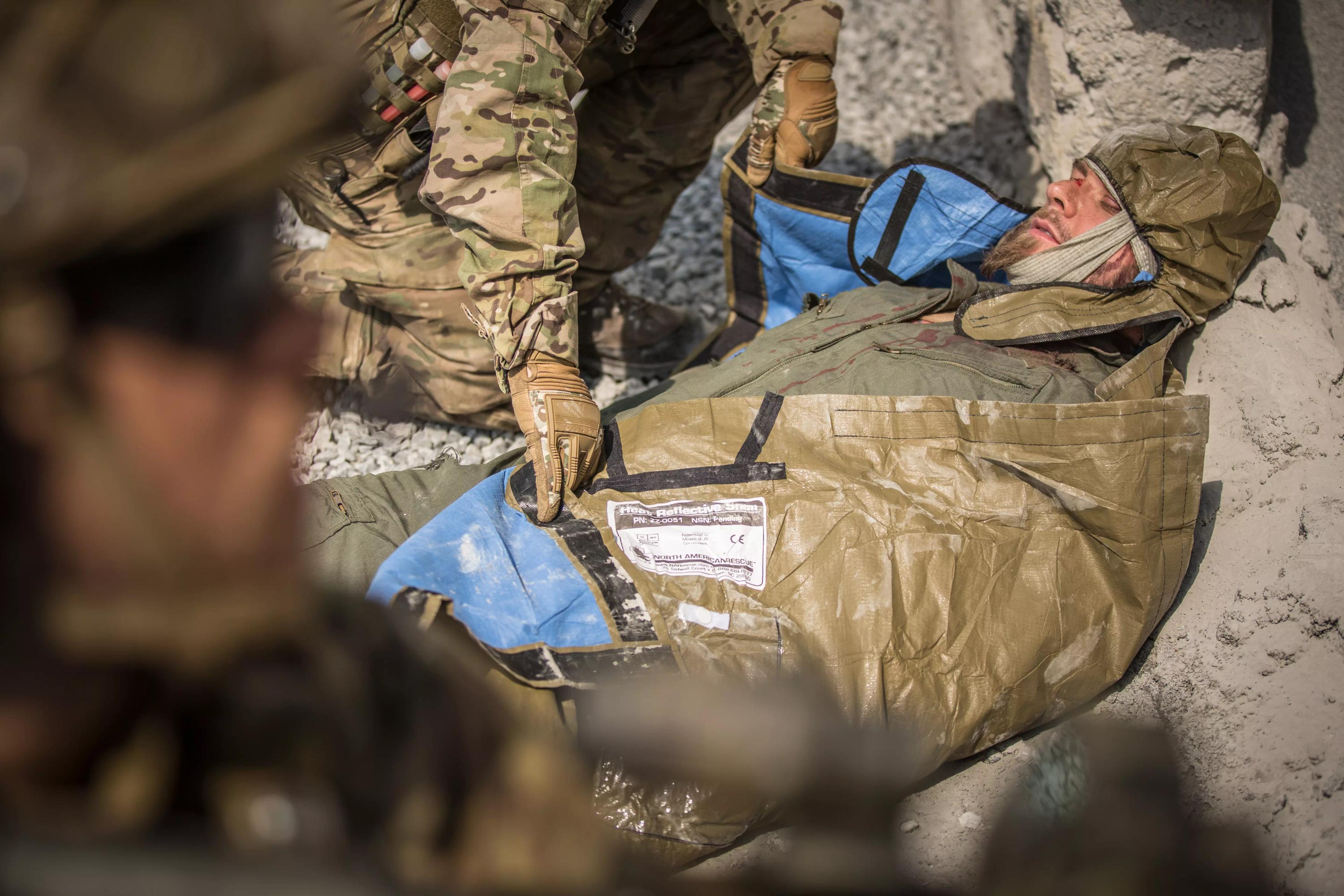 Ein Soldat kniet und versorgt einen mit dem NAR Hypothermia Prevention and Management Kit (HPMK®) geschützten Kameraden auf einer blauen Notfallplane.