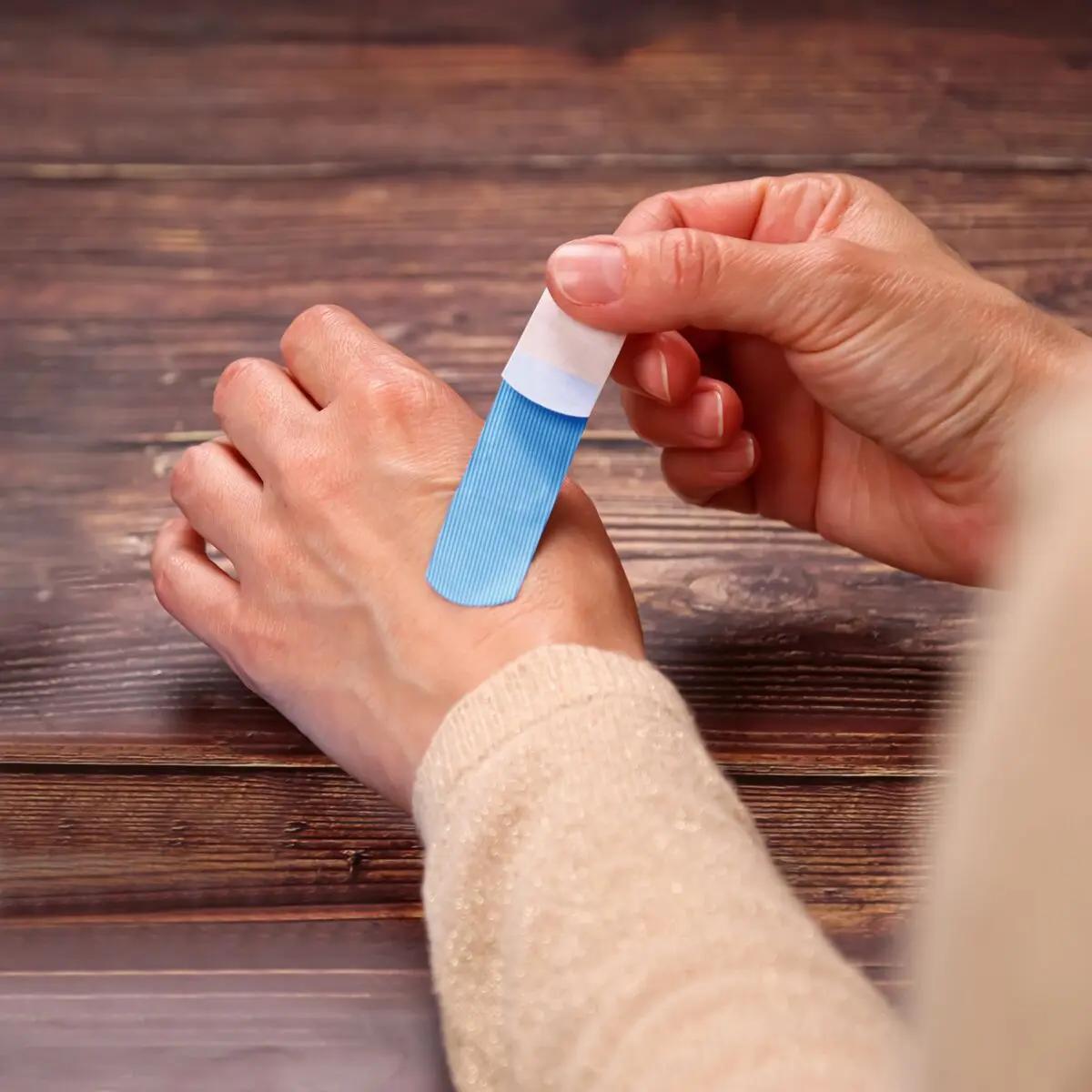 Eine Hand hält einen blauen Werosmart® AquaFlex Detect Pflasterstrip mit weißem Abziehstreifen über zwei Finger, das Pflaster ist wasserdicht und detektierbar.