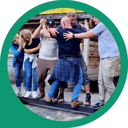 Sechs Personen balancieren und unterstützen sich gemeinsam auf einer Holzplanke.