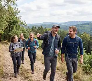 Vier Personen wandern mit Rucksäcken und Karte auf einem Waldweg durch bewaldetes Hügelland unter teils bewölktem Himmel.