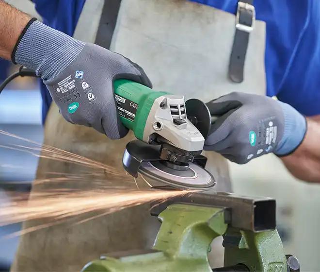 Person mit Schutzhandschuhen arbeitet mit grün-silbernem Winkelschleifer an Metall, Funkenflug beim Schleifen, grüner Amboss im Vordergrund.