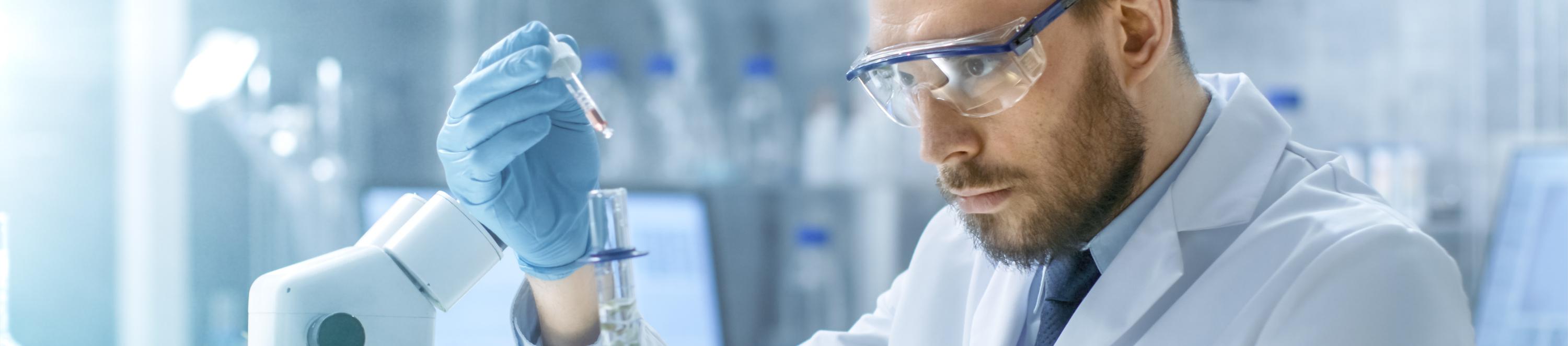 Ein Laborant mit Kittel und Schutzbrille füllt eine Flüssigkeit mit einer Pipette in ein Reagenzglas.