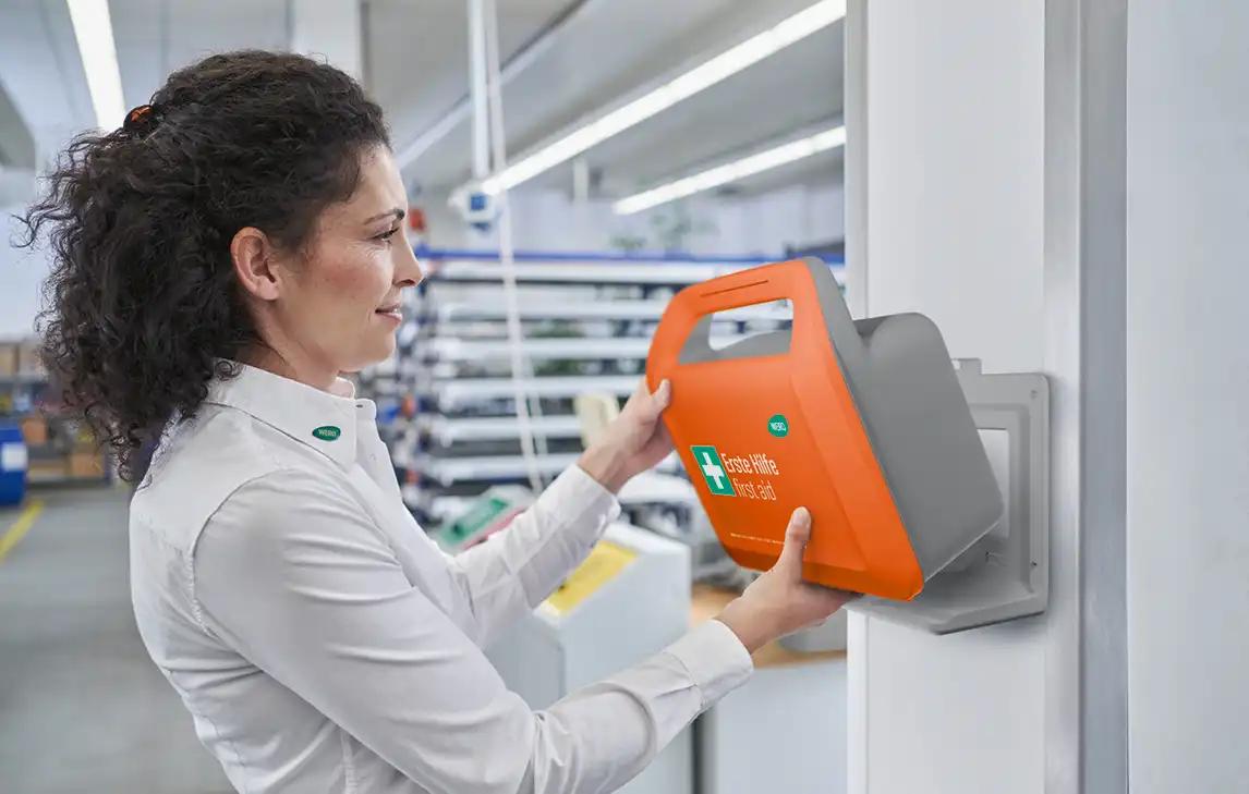 Eine Frau mit dunklen Locken nutzt ein orange-graues Erste Hilfe Gerät mit grünem Logo und dem Schriftzug „Erste Lufe First Aid“.