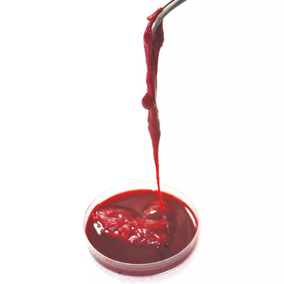Eine kleine Petrischale mit TrueClot® Gerinnungslösung, tiefrot und viskos, wird von einer Metallpipette zum Tropfen gebracht.