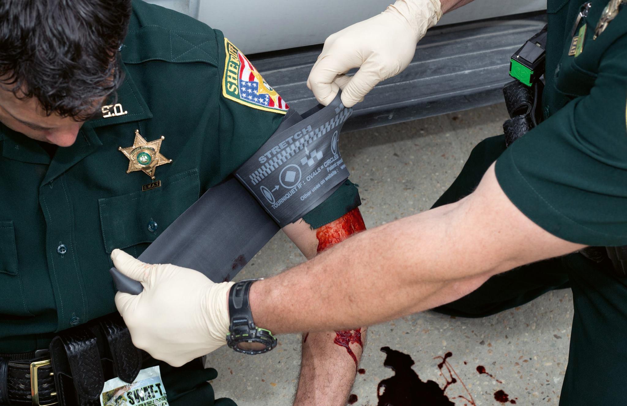Polizist legt sich mithilfe eines Tourniquet SWAT - T - Schwarz mit Unterstützung sterile schwarze Binde an verletztem, blutendem Arm an.