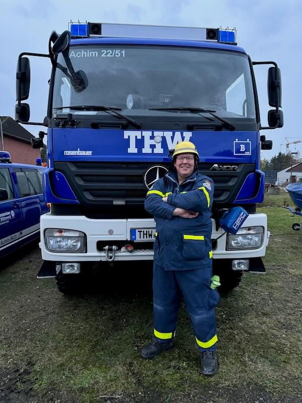 Ein Feuerwehrmann in blauer und gelber Schutzkleidung mit Helm steht lächelnd vor einem blauen THW-Löschfahrzeug mit der Aufschrift "Achim 22/51".