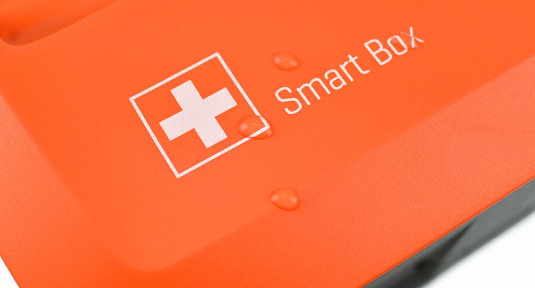 Orangefarbene Kunststoffbox mit weißem Kreuz und Aufschrift „Smart Box“, zwei Wassertropfen auf der Oberfläche sichtbar.