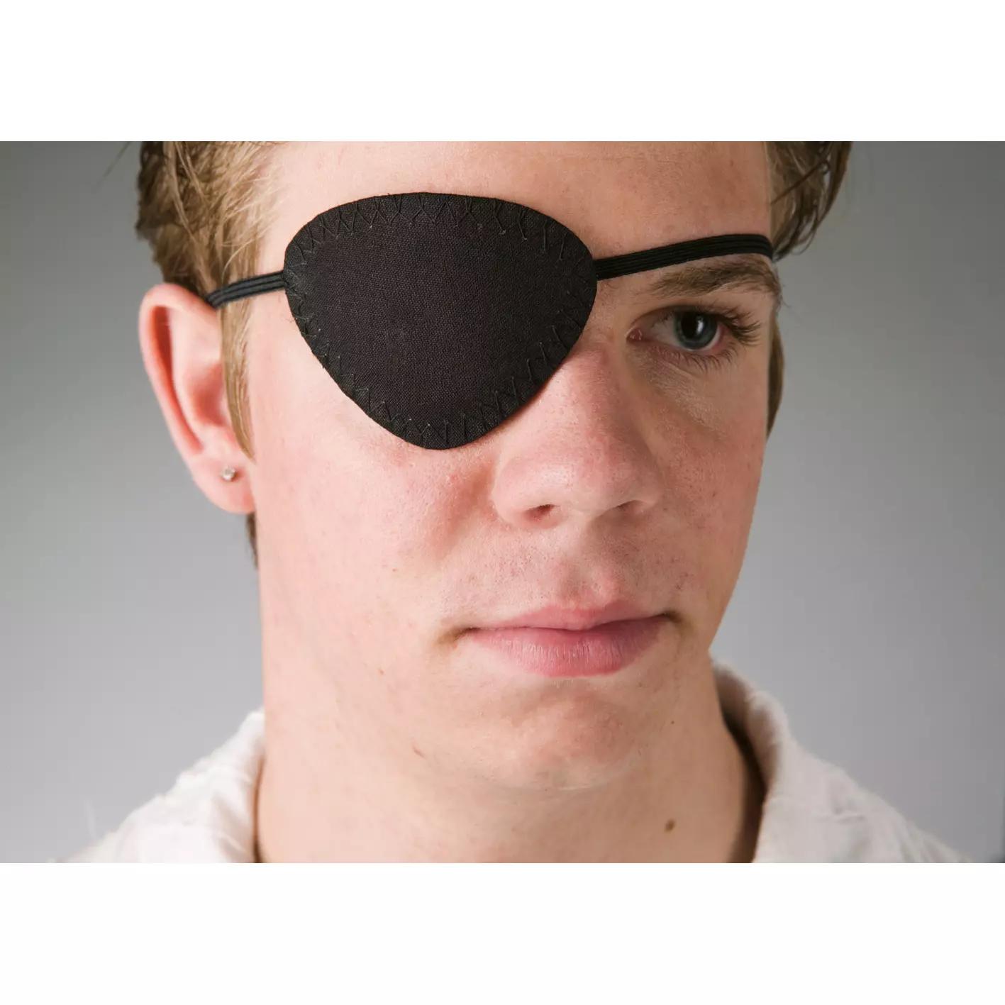 Ein junger Mann mit blonden Haaren trägt eine ovale Augenklappe aus schwarzer Baumwolle mit flexiblem Befestigungsband über dem linken Auge.