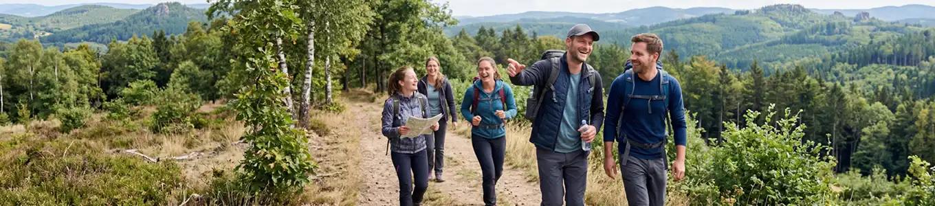 Fünf Menschen mit Rucksäcken wandern auf einem Waldweg, umgeben von Bäumen und hügeliger Landschaft im Hintergrund.