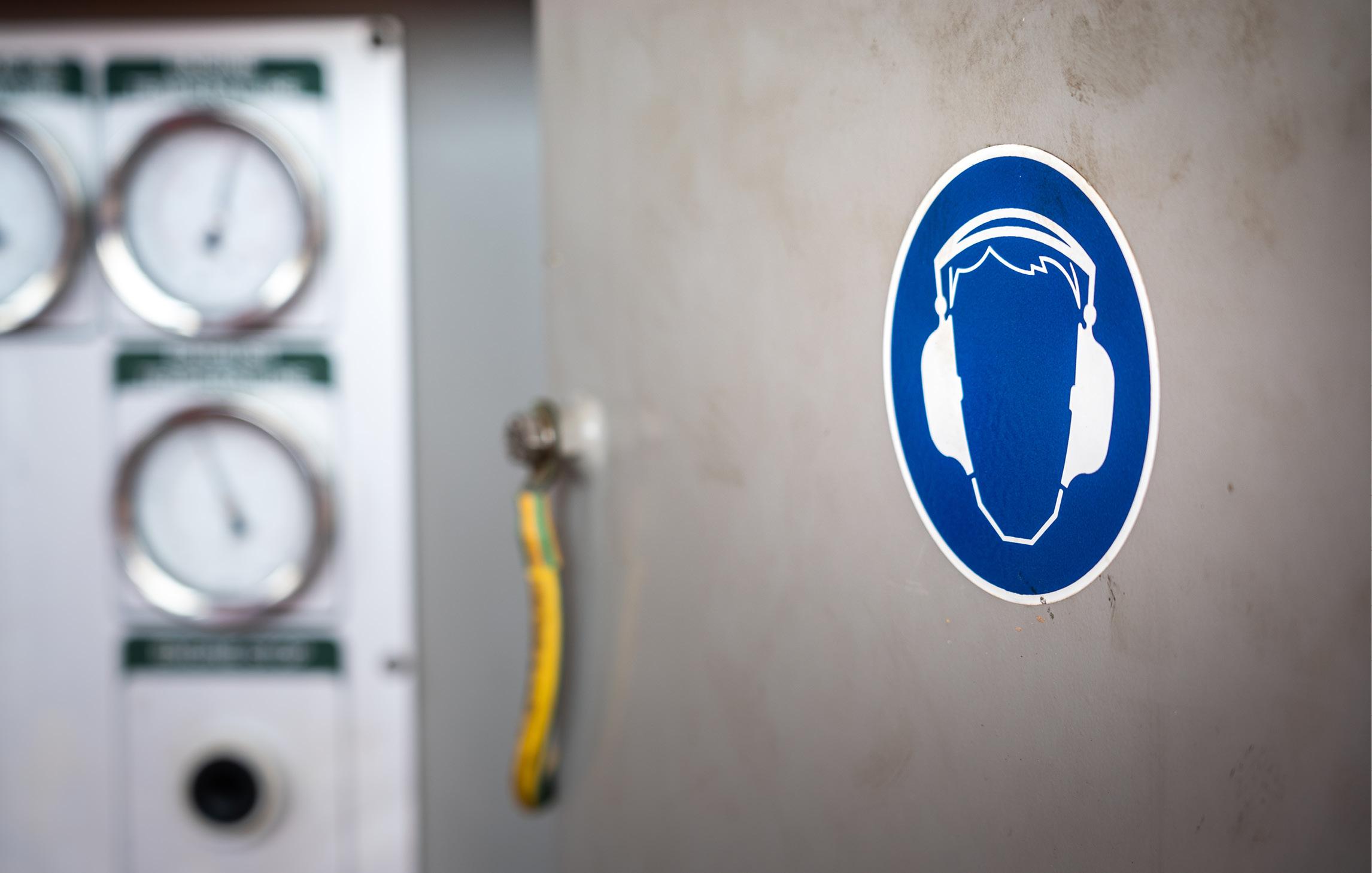 Im Hintergrund sind verschiedene Apparaturen zur Messung erkennbar. Im Vordergrund ist eine Wand in Betonoptik mit dem Hinweiszeichen "Gehörschutz"