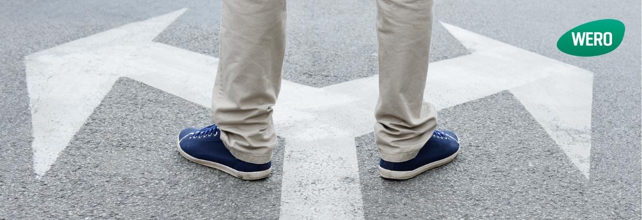Eine Person in beige Hosen und blauen Sneakers steht an einer Kreuzung mit zwei abzweigenden Wegen, die durch weiße Pfeile auf grauem Asphalt gekennzeichnet sind.