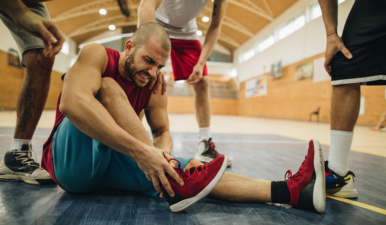 Ein Basketballspieler sitzt verletzt auf dem Hallenboden, fasst sein rechtes Bein, während zwei Teamkameraden ihm unterstützend beistehen.