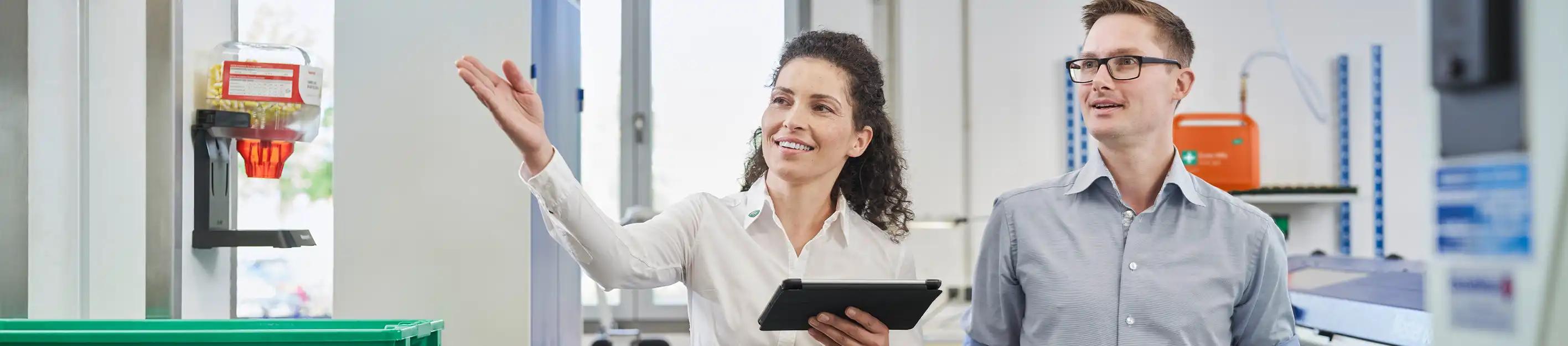 Eine Frau mit Tablet spricht gestikulierend mit einem Mann im Labor, im Hintergrund Laborgeräte und Regale.