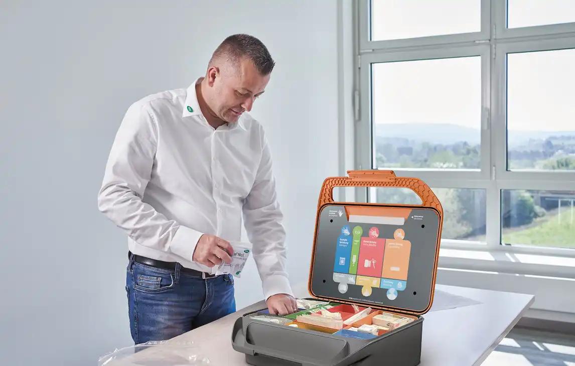 Ein Mann im weißen Hemd betrachtet eine geöffnete orange-schwarze Werkzeugbox mit Display und hält ein weißes Paket in der Hand.