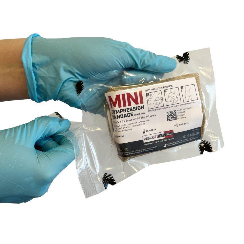 Die Rescue Essentials® Mini Compression Bandage wird in blauen Handschuhen gehalten, eingeschweißt in transparenter Verpackung, gut lesbar beschriftet und für Notfälle gekennzeichnet.