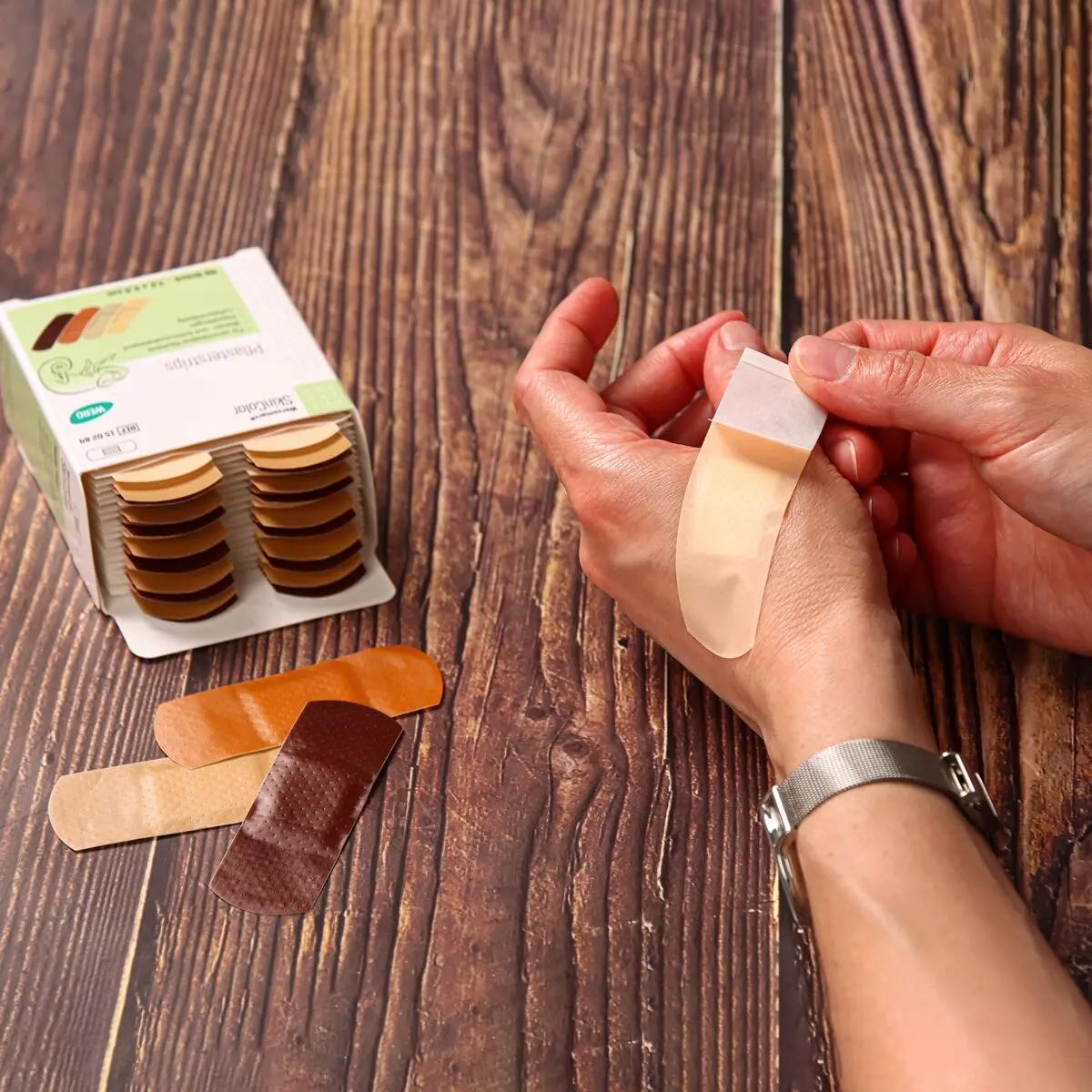 Eine Hand mit silberner Armbanduhr hält einen Werosmart® SkinColor Pflasterstrip, daneben liegen in verschiedenen Hauttönen weitere Pflaster und eine offene Kartonbox.