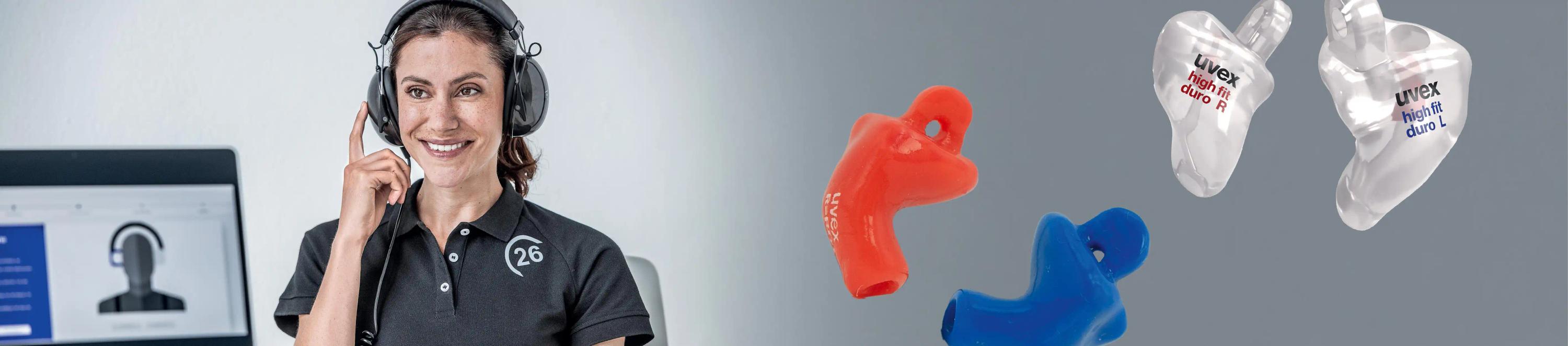 Eine Frau in schwarzem Poloshirt mit Kopfhörern sitzt an einem Schreibtisch und lächelt während eines Telefonats, ein Computer ist im Hintergrund sichtbar.