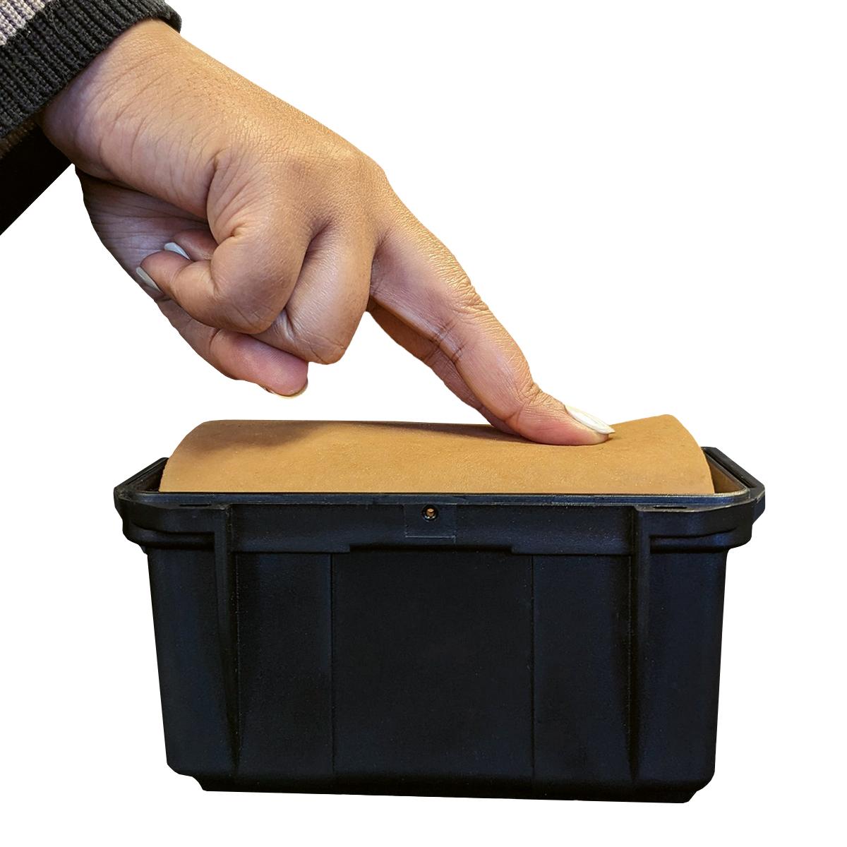 Eine Hand mit dunklem Ärmel berührt den Techline Entlastungspunktions-Trainer, ein schwarzes Kunststoffgehäuse mit simuliertem Haut- und Knochenmaterial.