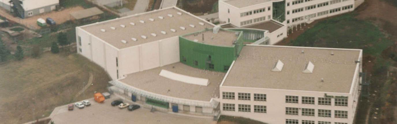 Großer Industriekomplex mit zentralem weiß-grünen Gebäude, umgeben von Nebenbauten, Parkplätzen, Fahrzeugen, Tanks und vereinzelten Grünflächen.