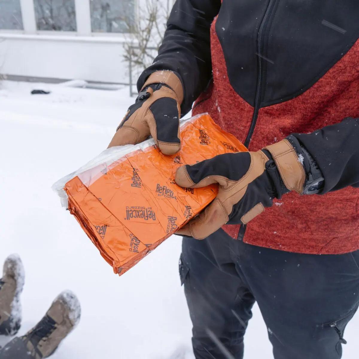 Eine Person hält einen in Folie verpackten Blizzard - Orange, Schlafsack mit Reflexcell™ Technologie und zellartiger Wärmeisolierung im Schnee.