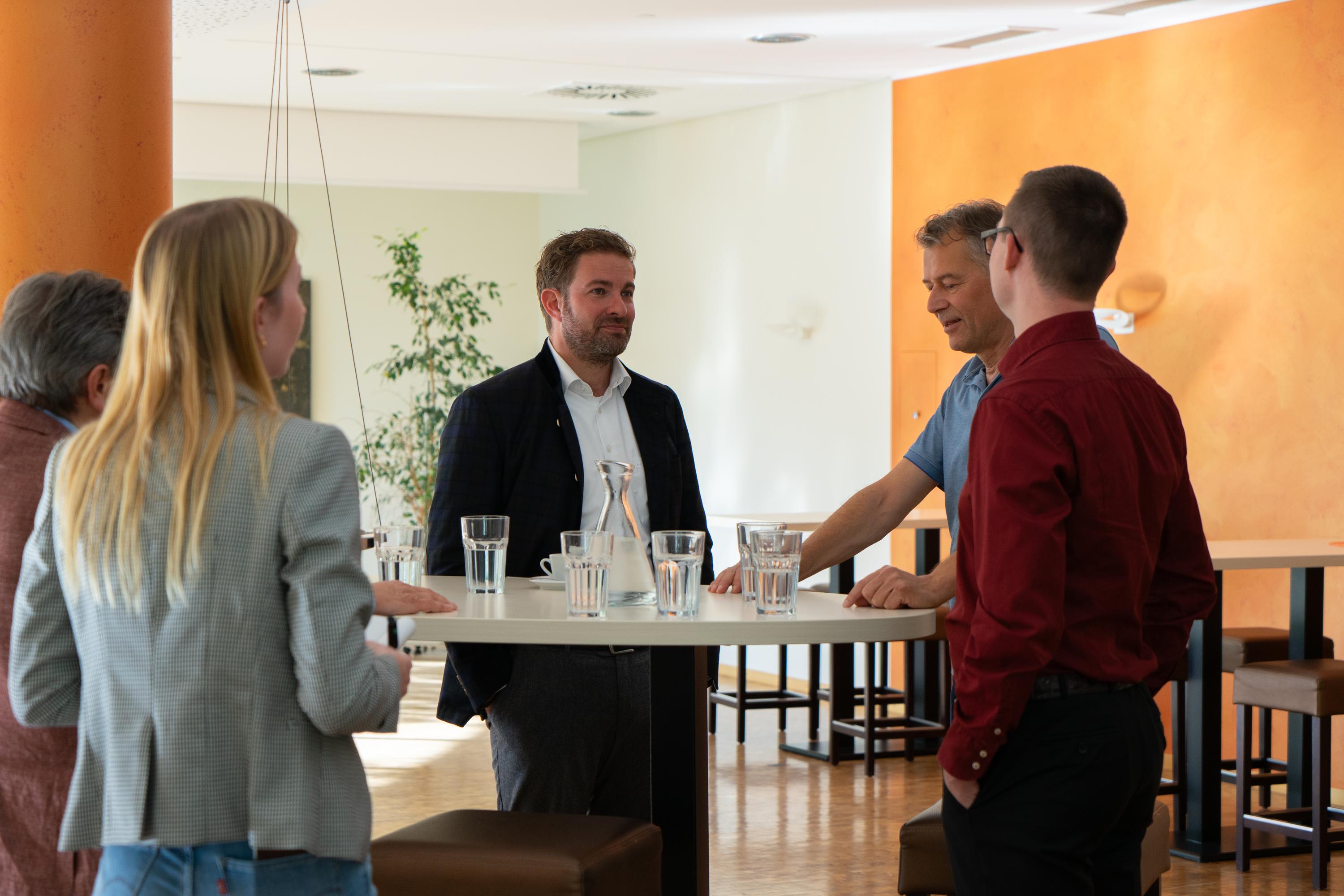 Fünf Personen sitzen an einem weißen Tisch im modernen Büro und führen ein Gespräch, umgeben von Wasserflaschen, Hockern und einer grünen Pflanze.