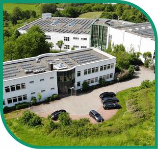 Großer weißer Gebäudekomplex mit Flachdach-Solaranlagen, angrenzendem Parkplatz mit schwarzen Autos und umgeben von grüner Vegetation.