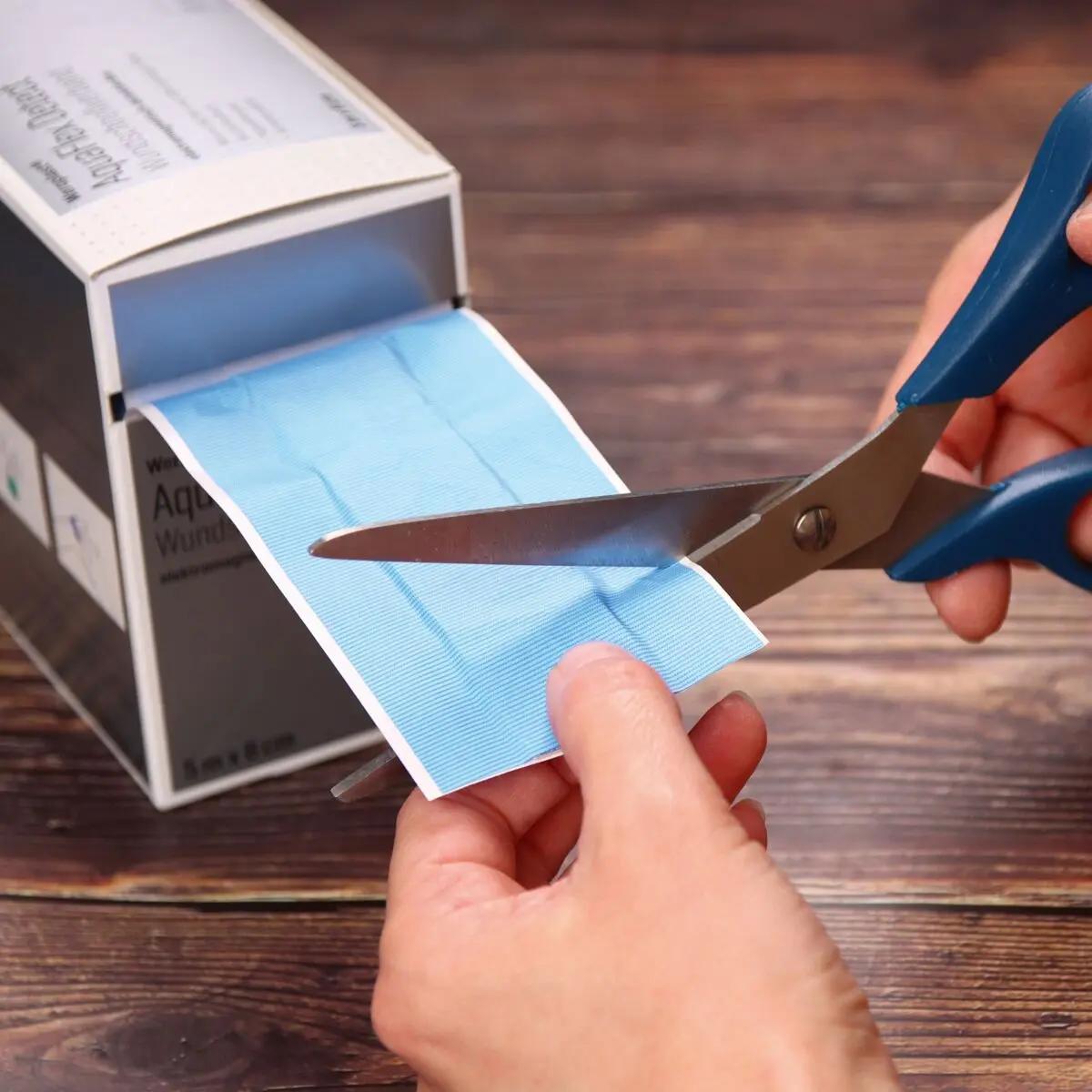 Eine blaue Schere schneidet einen Weroplast® AquaFlex Detect Wundschnellverband mit wasserdichter, blauer PU-Trägerfolie aus einer Box.