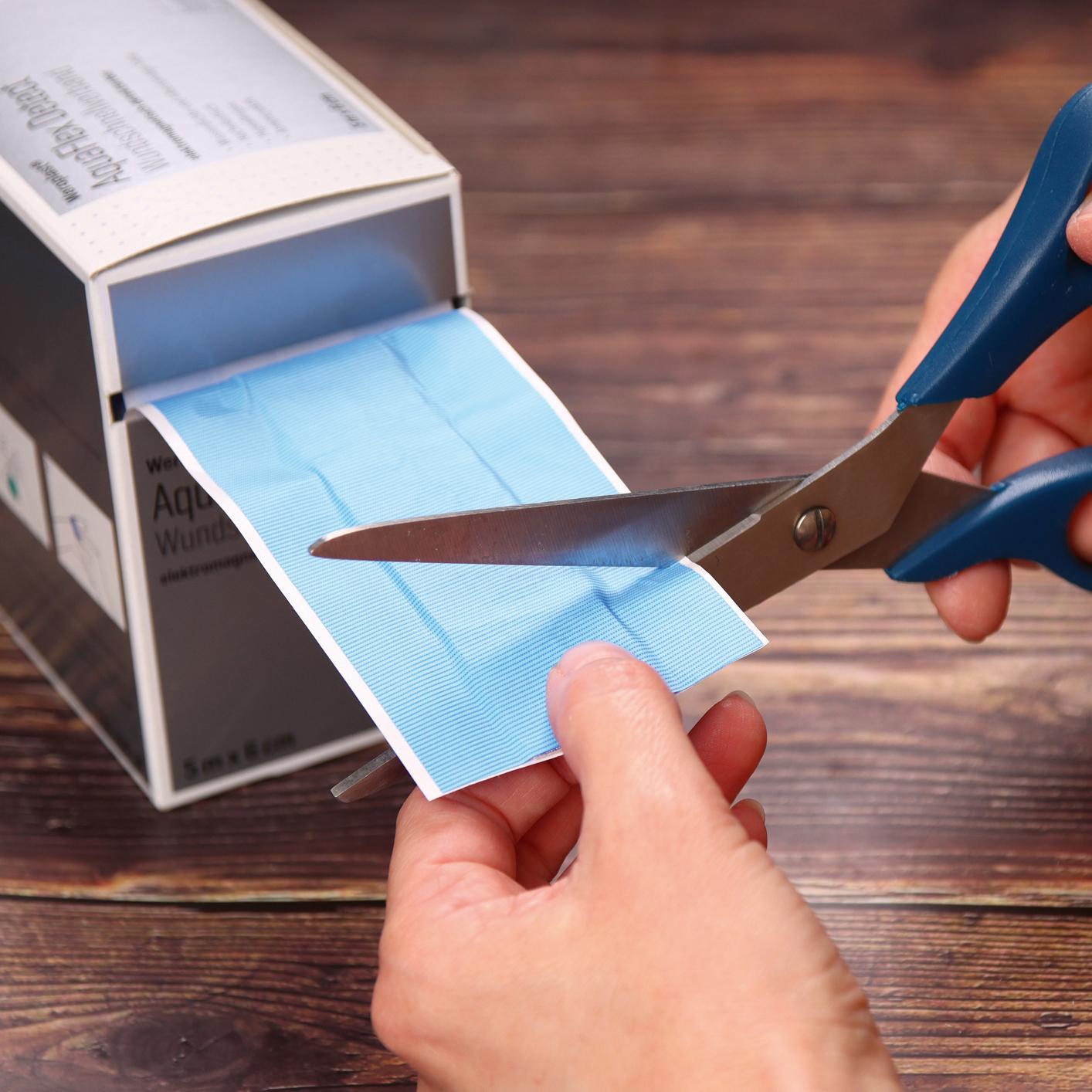 Eine blaue Schere schneidet einen Weroplast® AquaFlex Detect Wundschnellverband mit wasserdichter, blauer PU-Trägerfolie aus einer Box.