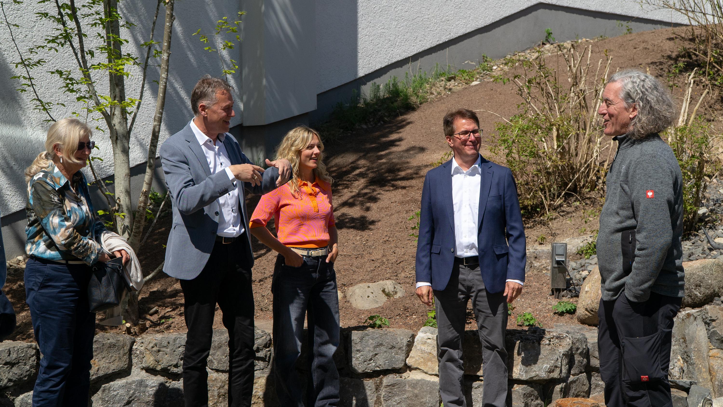 Fünf Personen mittleren Alters unterhalten sich im Garten vor einem weißen Gebäude, eine Frau zeigt in eine Richtung, eine andere hält ein Handtuch.