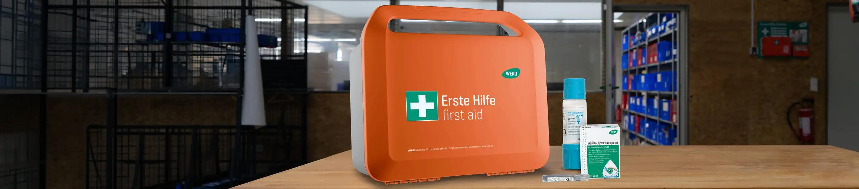 Orangefarbener Erste-Hilfe-Kasten mit Griff, daneben Wundcreme, grüne Erste-Hilfe-Schachtel und weiße Box auf Holztisch.