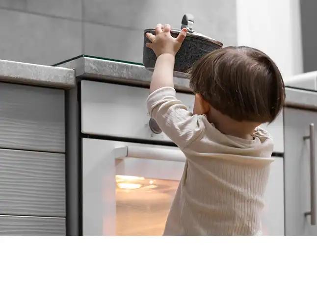 Kleines Kind im gelben Shirt steht mit ausgestreckten Armen vor einem offenen Ofen in einer modernen Küche.