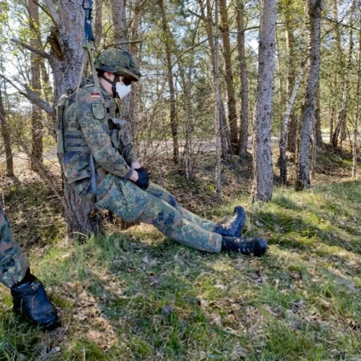 Ein Soldat sitzt am Boden, gesichert mit der TREMA Rescue Sling (TRS) um Oberkörper und Hals, schwarze Handschuhe und Stiefel tragend, von Seilen gehalten.