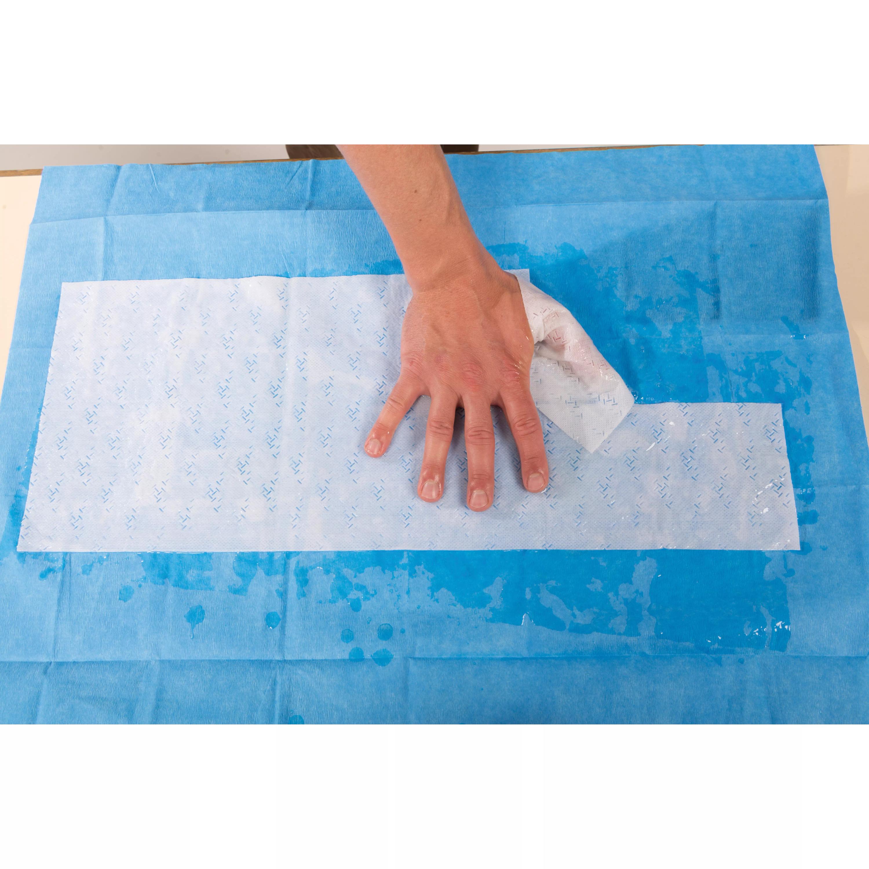 Eine Hand wischt blaue Flüssigkeit von einer glatten Oberfläche mit der WATER-JEL® HA First Responder Handkompresse, steril, 20 x 55 cm.
