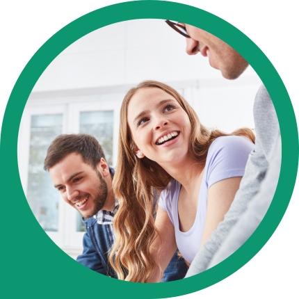 Eine lachende blonde Frau in Lila, ein Mann mit Bart und ein Mann mit Brille blicken glücklich in die Kamera.