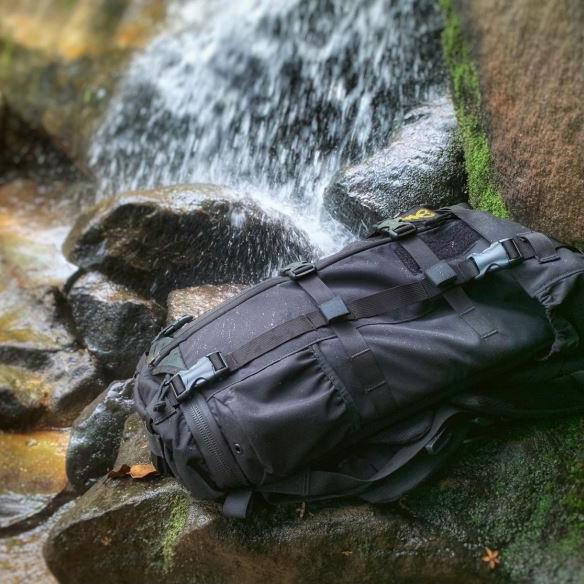 Schwarzer Rucksack mit mehreren Riemen liegt auf einem braunen Felsen neben einem Wasserfall inmitten grüner Natur.