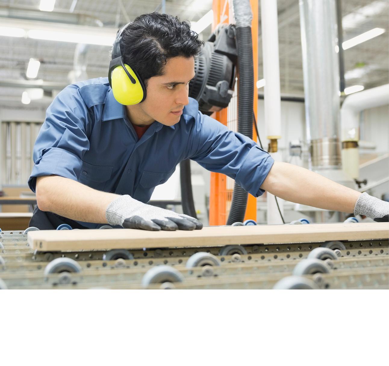 Mann steht an einer Sägemaschine und passt ein Stück Holz an. Er trägt Arbeitshandschuhe und gelbe Kapselgehörschützer