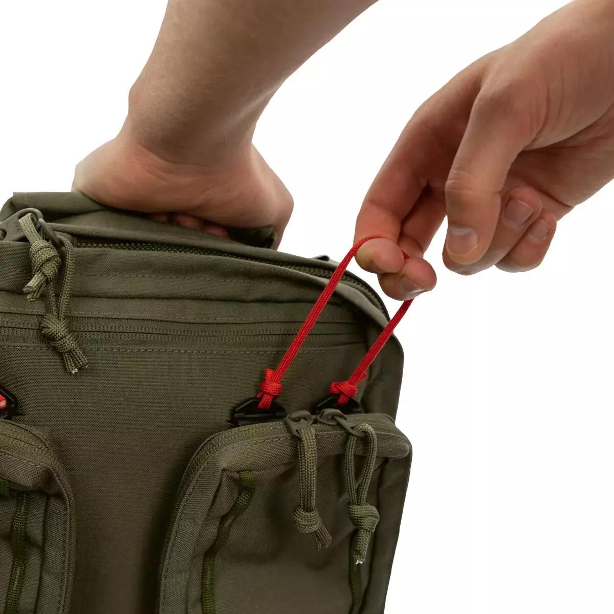 Eine Hand hält einen roten Kordelzug am TT Medic Mascal Pack - Oliv, der mehrere Frontfächer mit Reißverschluss für schnellen Zugriff hat.