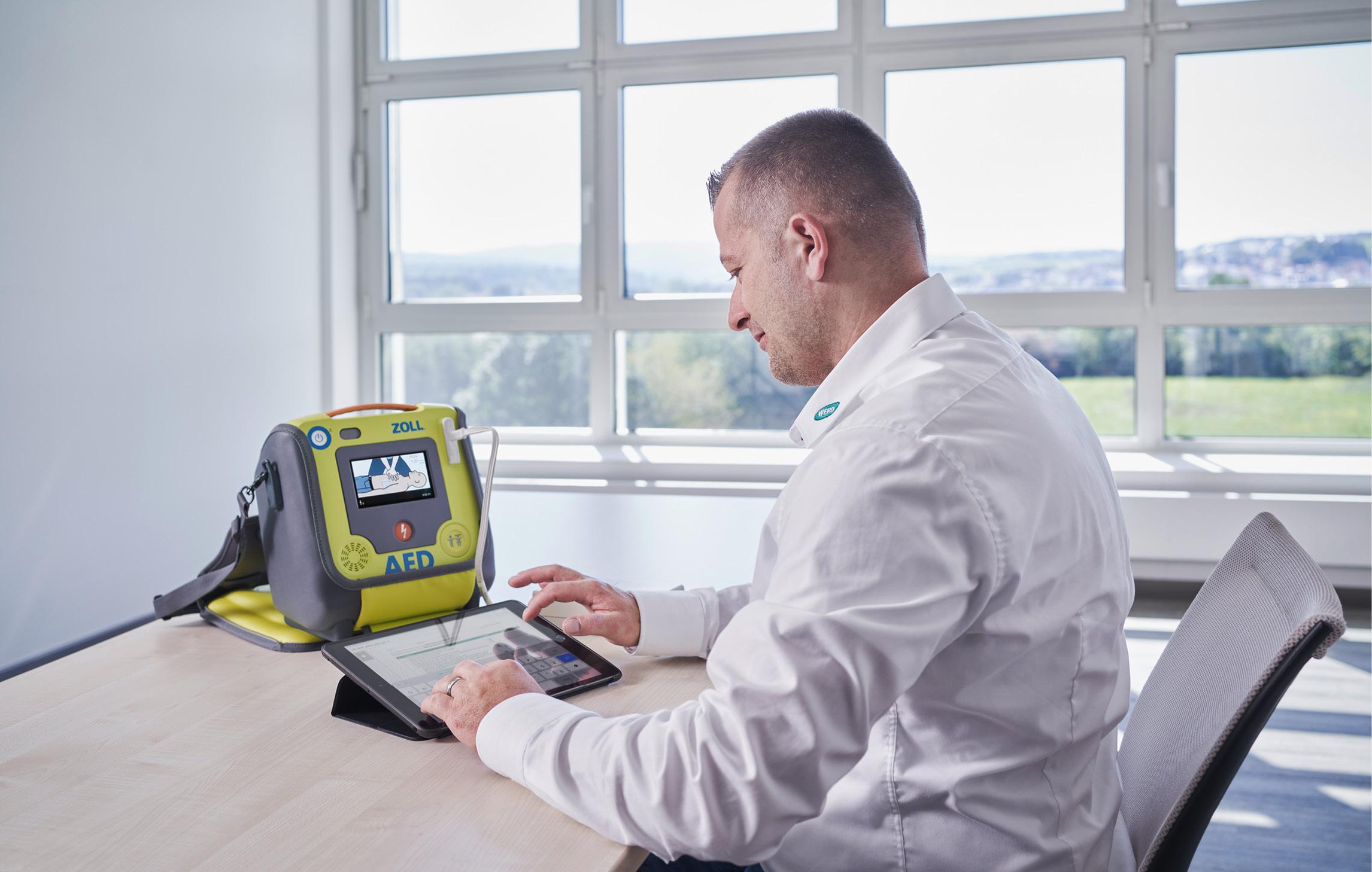 Ein WERO Mitarbeitender sitzt an einem Tisch und prüft über sein iPad die Funktionen eines vor ihm stehenden AED Defibrillators