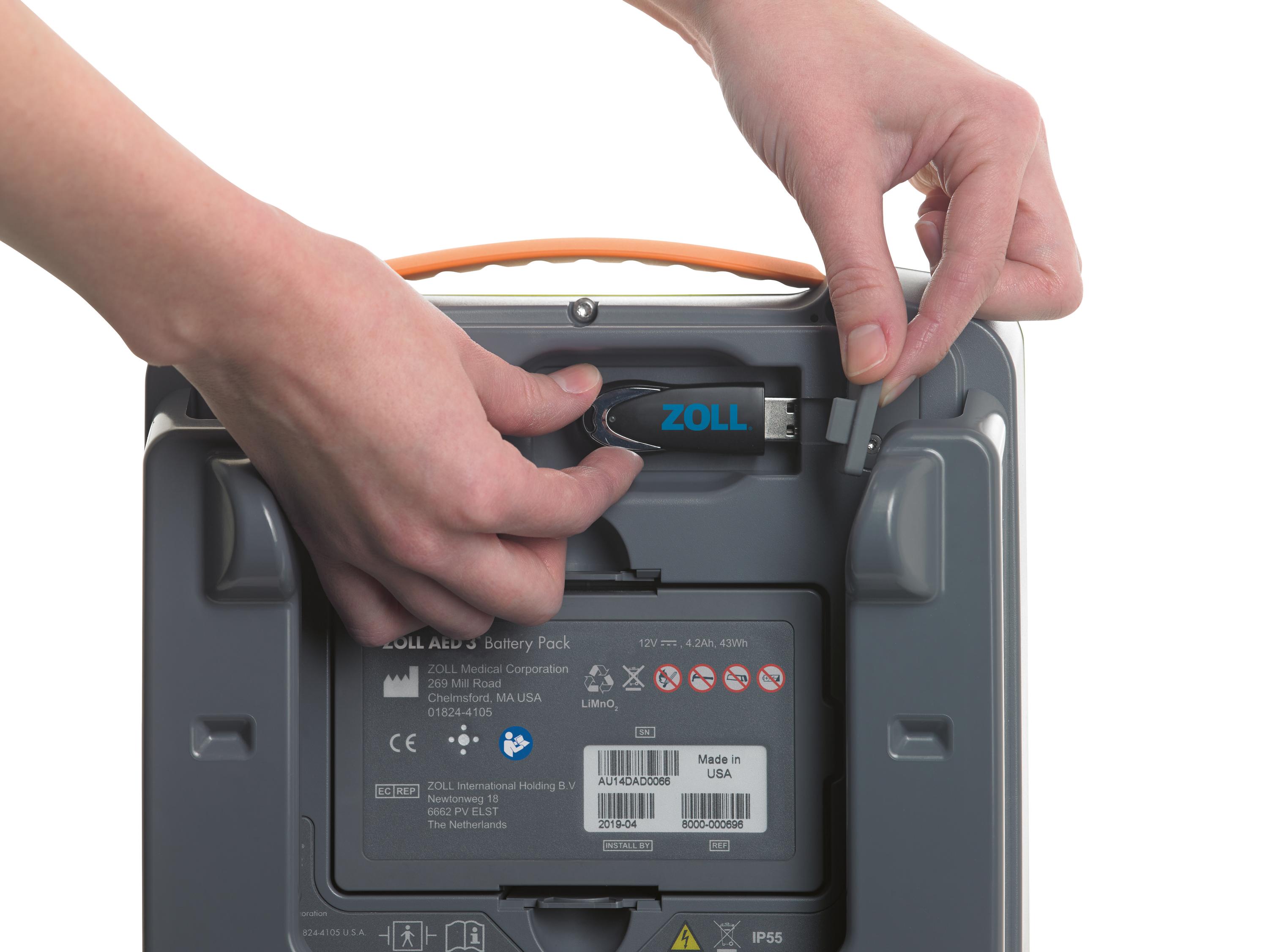 Ein Zoll AED 3 Batteriepack wird von zwei Händen gehalten, sichtbar sind die angeschlossenen Batterien, das orangefarbene Griffstück und ein Zoll USB-Kabel.