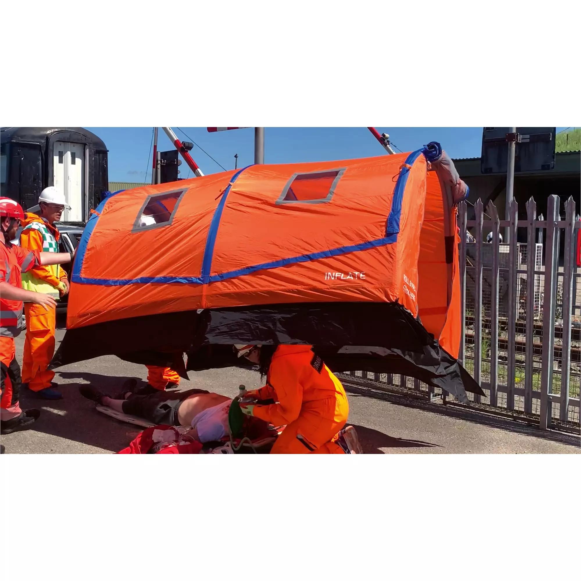 Mehrere Helfer in orangener Schutzkleidung versorgen eine Person auf einer Trage und bringen ein aufgeblasenes RIGLOO Rescuer Rettungszelt darüber an.