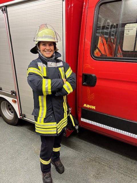 Feuerwehrmann in gelber Schutzkleidung mit Reflexstreifen steht lächelnd, Arme verschränkt, neben rotem Feuerwehrfahrzeug mit „ADIK“-Aufschrift.