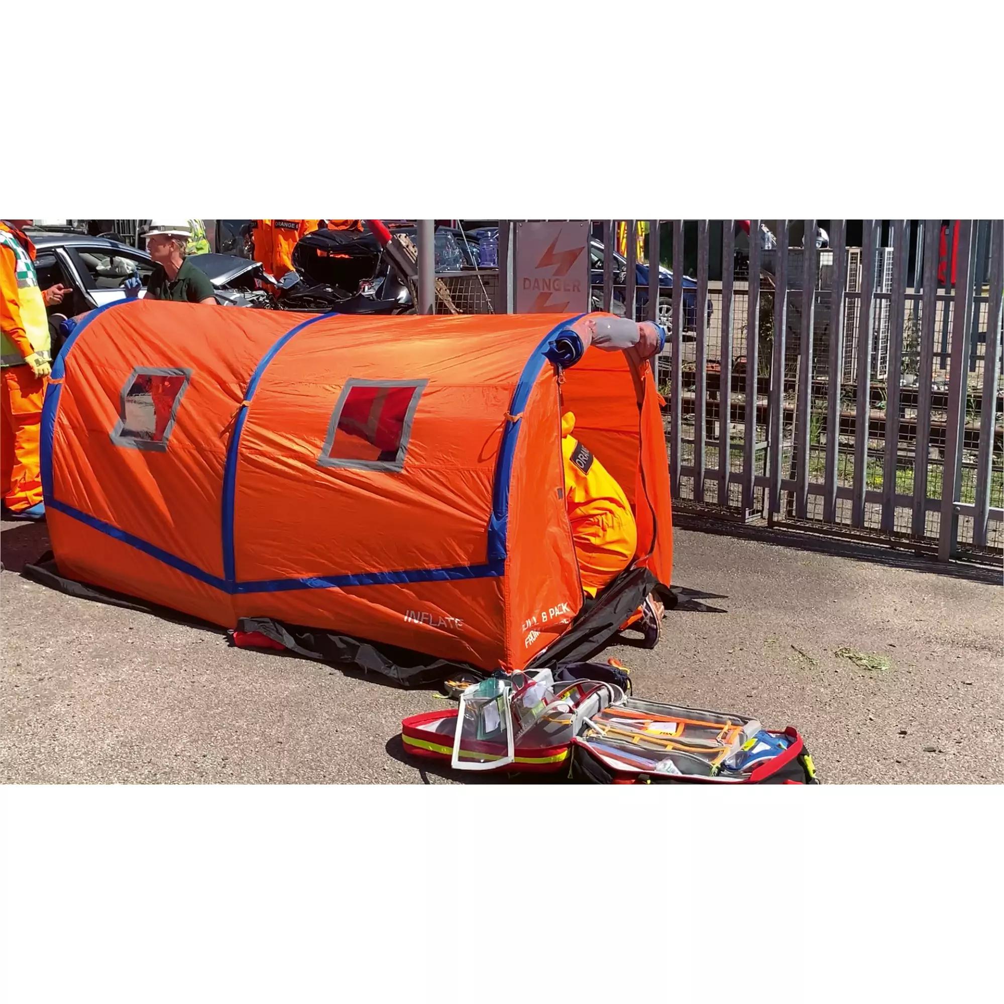 Ein orange-blaues RIGLOO Rescuer Rettungszelt mit mehreren Fenstern liegt teils aufgeblasen am Boden, daneben Rettungsausrüstung und zwei Einsatzkräfte.